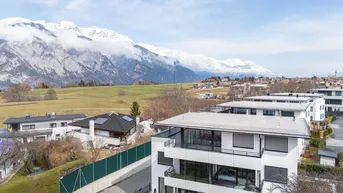 Moderne Wohngebäude mit Flachdach und Balkonen, umgeben von grünen Feldern und schneebedeckten Bergen.