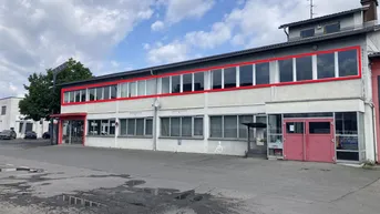 Gebäudeansicht mit großen Fenstern im Obergeschoss und Parkplatz davor unter bewölktem Himmel.
