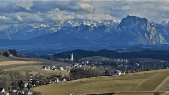 Panoramablick auf ein Dorf in einem Tal mit schneebedeckten Bergen im Hintergrund und weiten Feldern.