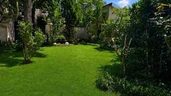 Gepflegter Garten mit sattgrünem Rasen, umgeben von Bäumen und Sträuchern unter blauem Himmel.