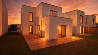 Modernes Einfamilienhaus mit Holzterrasse und Balkon, beleuchtet vom warmen Licht des Sonnenuntergangs.