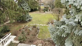 Weitläufiger Garten mit Rasenfläche, Zierbeeten und altem Baumbestand, von einer Terrasse aus gesehen.