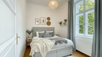 Helles Schlafzimmer mit gemütlichem Bett, Parkettboden und großem Fenster mit Blick ins Grüne.