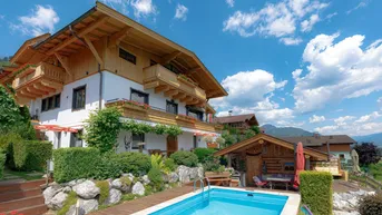 Charmantes Gebäude mit Holzbalkonen, umgeben von einem gepflegten Garten und einem erfrischenden Pool unter blauem Himmel.