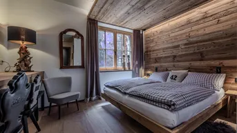Gemütliches Schlafzimmer mit Holzwand, Holzbalkendecke und einem Fenster mit Blick ins Grüne.