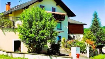 Ansicht des Hauses von außen mit Balkon, umgeben von viel Grün und Bäumen unter blauem Himmel.