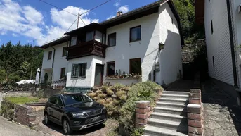 Weißes Wohnhaus mit Balkon und Außentreppe, umgeben von grüner Vegetation unter blauem Himmel.