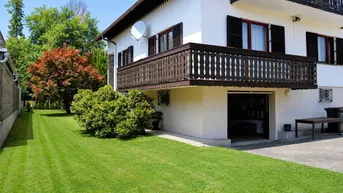 Weißes Haus mit Holzbalkon, gepflegtem Rasen und Bäumen im Gartenbereich.