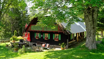 Traditionelles Holzhaus mit Balkon und gemütlichem Außenbereich, umgeben von grüner Natur und Bäumen.