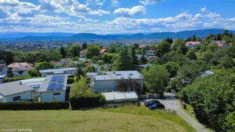 Luftaufnahme einer Wohnsiedlung mit Häusern, viel Grün und einem weiten Blick auf die Stadt und die Berge.