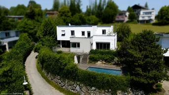 Modernes weißes Haus mit Flachdach, umgeben von viel Grün, inklusive Außenpool und Zufahrt.