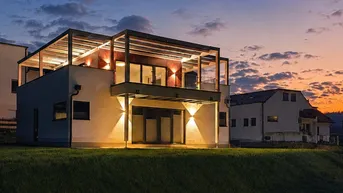 Modernes Haus bei Sonnenuntergang mit beleuchteter Fassade und Pergola auf dem Balkon.