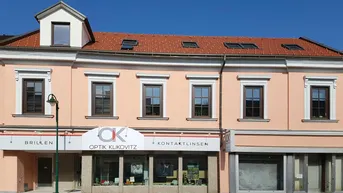 Fassade eines Gebäudes mit Geschäften im Erdgeschoss und Wohnungen darüber, unter blauem Himmel.