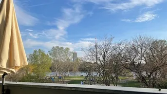 Panoramablick vom Balkon auf den See, umgeben von Bäumen und einem Spielplatz unter blauem Himmel.
