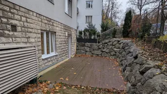 Kleine Holzterrasse am Gebäude, umgeben von einer Steinmauer und herbstlichem Laub.