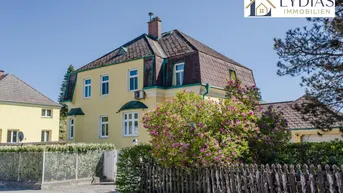 Historische Stadtvilla mit markantem Mansarddach, umgeben von einem Holzzaun und blühenden Büschen.
