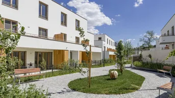 Moderner Wohnkomplex mit weißer Fassade, begrüntem Innenhof und gepflasterten Wegen unter blauem Himmel.