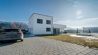 Modernes Einfamilienhaus mit gepflasterter Auffahrt, großem Garten und Trampolin unter blauem Himmel.