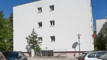Weißes Mehrfamilienhaus mit schlichter Fassade und Parkplätzen im Vordergrund unter blauem Himmel.