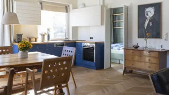 Offene Küche mit blauen Unterschränken, Holzarbeitsplatte und Fischgrätparkett, angrenzend an einen Schlafbereich.