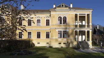 Historisches Gebäude mit klassischer Architektur und gepflegtem Vorgarten unter blauem Himmel.