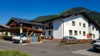 Traditionelles Gebäude im alpinen Stil mit weißer Fassade, Holzelementen und Carport in bergiger Umgebung.