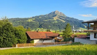 Haus mit rotem Ziegeldach und Holzzaun vor einer beeindruckenden Bergkulisse unter blauem Himmel.