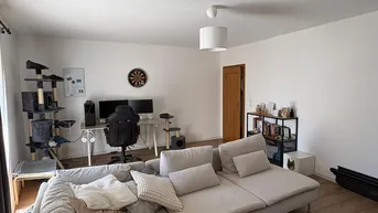 Geräumiges Wohnzimmer mit beigem Ecksofa, Holzboden und einem Arbeitsbereich mit zwei Monitoren.