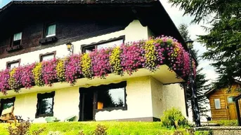 Traditionelles Haus mit blühendem Balkon und gepflegtem Gartenbereich unter blauem Himmel.