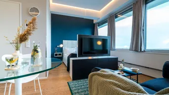 Modernes Studio-Apartment mit kombiniertem Wohn- und Schlafbereich, Esstisch und großen Fenstern.