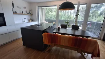 Moderne offene Küche mit dunkler Kücheninsel, Holzboden und Zugang zu einem Balkon mit Waldblick.
