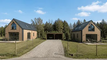 Zwei moderne Holzhäuser mit Satteldach, Kiesauffahrt und Carport in ländlicher Umgebung.