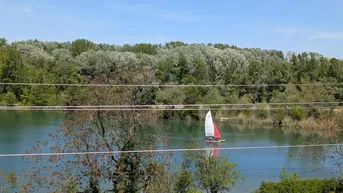 Blick auf einen malerischen See mit Segelboot, umgeben von grünen Bäumen unter blauem Himmel.