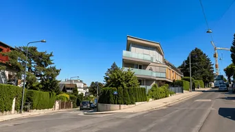 Moderne Architektur mit großen Fenstern und Balkonen an einer Wohnstraße, umgeben von viel Grün.