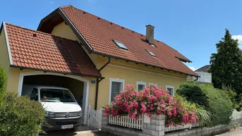Gelbes Einfamilienhaus mit rotem Ziegeldach, Garage und blühenden Sträuchern im Vorgarten.