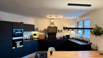 Moderne Küche mit schwarzen Fronten, integrierten Geräten und Holztisch, beleuchtet von stilvollen Hängelampen.