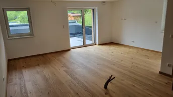 Helles, leeres Zimmer mit Holzboden und Zugang zu einer Terrasse, umgeben von grüner Natur.