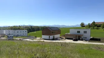 Neubaugebiet mit modernen Wohnhäusern und Mehrfamilienhäusern, umgeben von grünen Feldern und Bergpanorama.