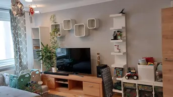 Helles Wohnzimmer mit TV-Möbel, Wandregalen und Blick ins Grüne durch große Fenster.