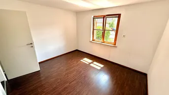 Helles Zimmer mit dunklem Laminatboden und einem Fenster mit Holzeinfassung.