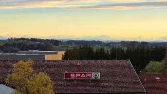Weitblick über Dächer und Bäume mit einem SPAR-Schild im Vordergrund und Bergen am Horizont.