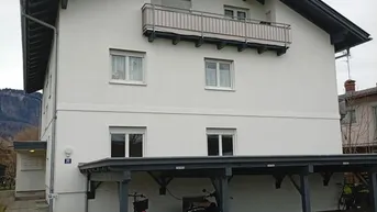 Weißes Mehrfamilienhaus mit dunklem Satteldach, Balkon und überdachtem Carport-Bereich.