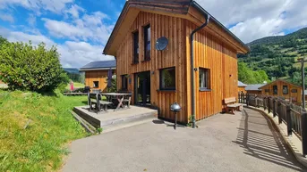 Modernes Holzhaus mit Terrasse und Bergblick unter blauem Himmel und viel Grün.