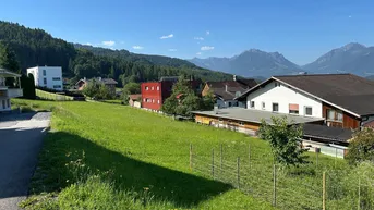 Weitläufige grüne Wiese mit umliegenden Häusern und beeindruckendem Bergblick unter klarem Himmel.
