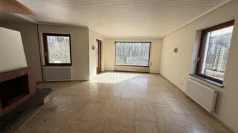 Leeres Wohnzimmer mit Fliesenboden, Kamin und großen Fenstern mit Blick ins Grüne.