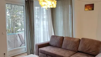 Gemütliches Wohnzimmer mit braunem Ecksofa, Teppich und Zugang zu einem kleinen Balkon.