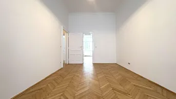 Helles Zimmer mit Fischgrätparkett, hohen Decken und Zugang zu weiteren Räumen.