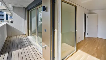 Heller Innenraum mit Holzboden und Zugang zu einem Balkon mit Holzbelag und Blick auf umliegende Gebäude.