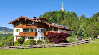 Traditionelles Haus im alpenländischen Stil mit Holzbalkonen, grünen Fensterläden und Solaranlage auf dem Dach.