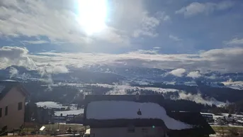 Panoramablick auf schneebedeckte Berge und ein Haus mit verschneitem Dach unter strahlender Sonne.